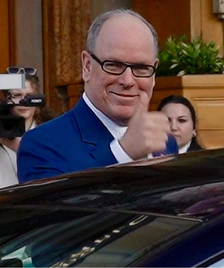 Fürst Albert steht in einem blauen Anzug und zeigt Daumen hoch, im Hintergrund stehen Menschen und eine Gebäude-Fassade.