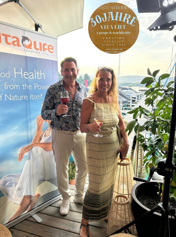 Zwei Personen halten Gläser vor einem Vita-Life Hintergrund, dahinter Wasser und Boote.