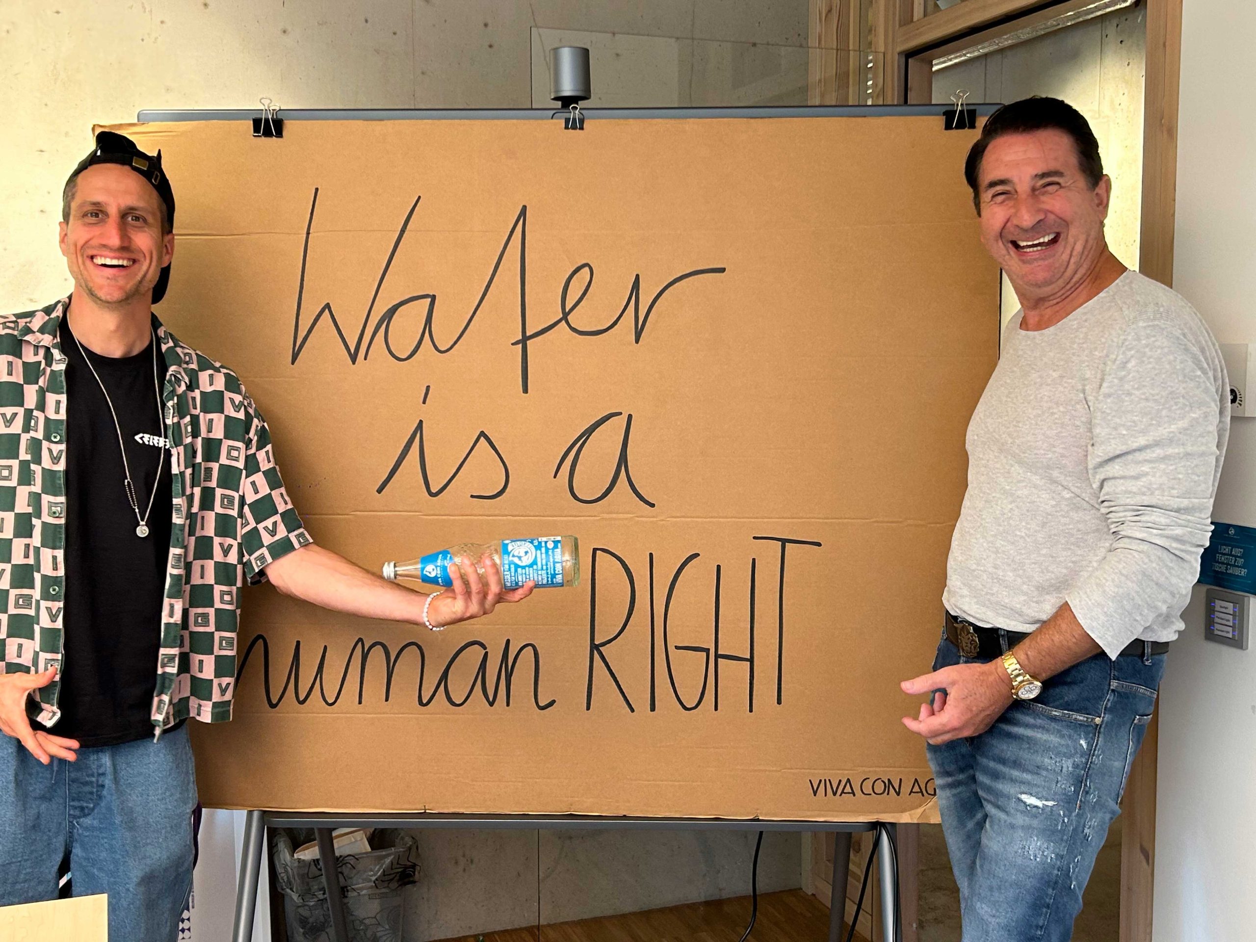 Mario Hintermayer steht mit einem Mann vor einem großen Schild „Water is a human right“, beide lachen, links wird eine Wasserflasche gezeigt, unten steht „Viva con Agua“.